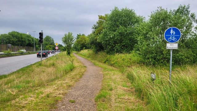 Farbfoto: Neben einer großen Straße mit zwei Fahrstreifen pro Richtung führt rechts ein  schmaler Weg mit Kies-/Schotteroberfläche entlang, beschildert als gemeinsamer Geh-/Radweg mit Zusatzschild "Kein Winterdienst". 