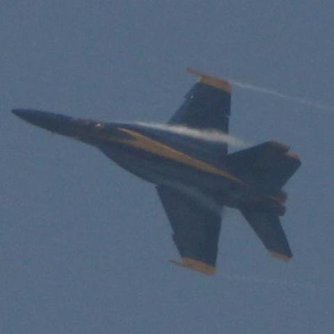 A hazy photo of a Blue Angels jet, in flight. This blue and gold United States Navy F/A-18 Super Hornet makes a hard banking turn. Condensation trails from it's engine intakes and wingtips.
