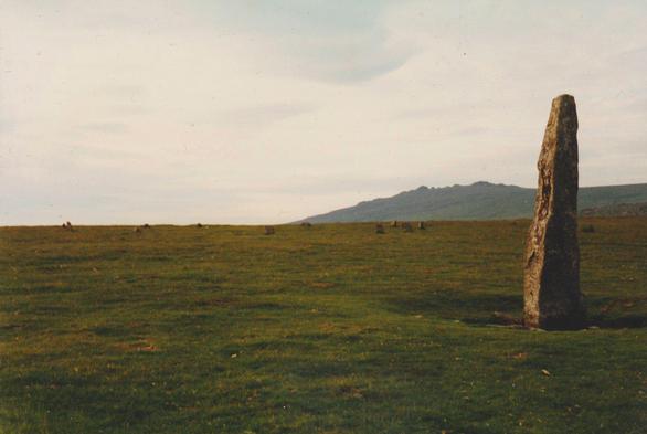 Colour photograph showing, on the right, the 3.1m tall menhir or prehistoric standing stone at Merrivale, between Princetown and Tavistock, on Dartmoor in Devon. On the left, in the middle distance, to the north of the menhir, is a stone circle, about 20.5 by 17.8m in circumference. Rising in the background can be seen Great Mis Tor.