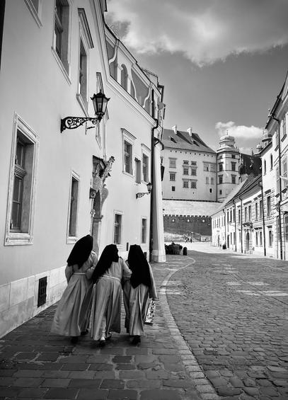Black and white photograph showing three nuns walking together holding each other under the arm. They are walking down the oldest street in Krakow, Poland. On both sides of the cobblestone street there are old tenement houses of different heights. The street is closed by a fragment of a building that is part of the Wawel Royal Castle. The Sun had just hidden behind a cloud trying to illuminate the entire scenery. 