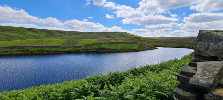 A narrow reservoir surrounded by moorland. It curves away in the distance so the end is hidden by the hill on the opposite bank. In the foreground, the bank is covered with ferns and there is a stone wall in a state of disrepair on the right. The sky is blue with over exposed white clouds.