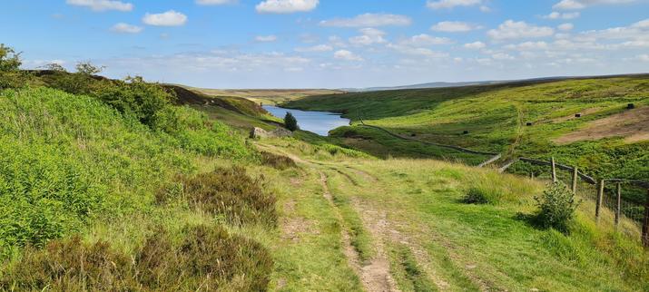 A view down a shallow valley, with green moorland hills on either side. At the far end of the valley is a reservoir. It's a bright sunny day, making the greenery and blue sky pop. A footworn path leads from the centre of the image in the direction of the far reservoir.