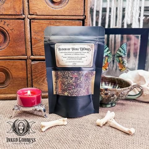 A pouch of loose leaf tea stands in front of a glazed earthenware teacup on a burlap-covered surface. There are bones scattered in front of the pouch and a red tealight for scale. The pouch has a clear window on the front with a label that features the name of the tea, "Blood of Thine Enemies," ingredients, and preparation instructions. This is an exclusive product from Inked Goddess Creations.