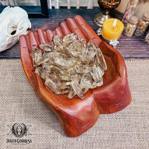 A variety of African Citrine Tumbled Point crystals rest in cupped, carved wooden hands. The crystals are elongated, smooth, and transparent with a pale honey color. Presented by Inked Goddess Creations.