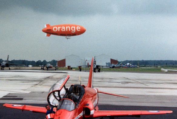 Orange blimp against a somewhat overcast sky. In the foreground can beseen one of the Hawks of the Red Arrows with its canopy open. In the middle distance a selection of aircraft, and in the background a buildings and trees.