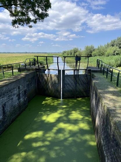 Sluice with green water in the shade, brilliant sunshine on the canal behind.