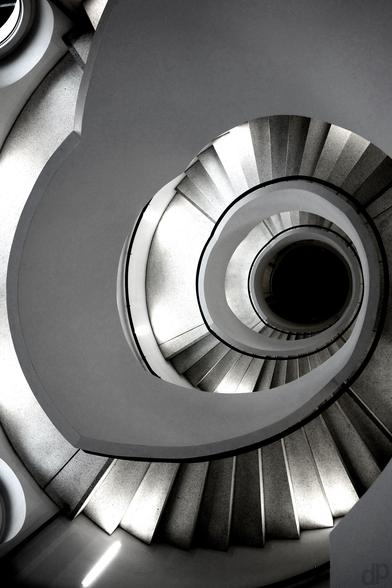 Schwarzweißaufnahme

Ein rundes Treppenhaus von oben senkrecht nach unten aufgenommen. Der Kontrast lässt manche Stufen weiß bzw. heller und manche sehr dunkel erscheinen.
Ein schwarzes Geländer zieht sich and er Treppeninnenseite bis nach unten durch. Alles verliert sich in einem schwarzen Kreis.