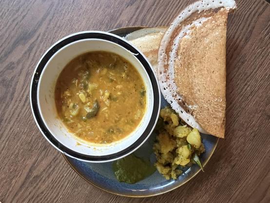 A bowl of sambher, a crispy dosa, potato curry, and a small portion of green chuney on a plate.