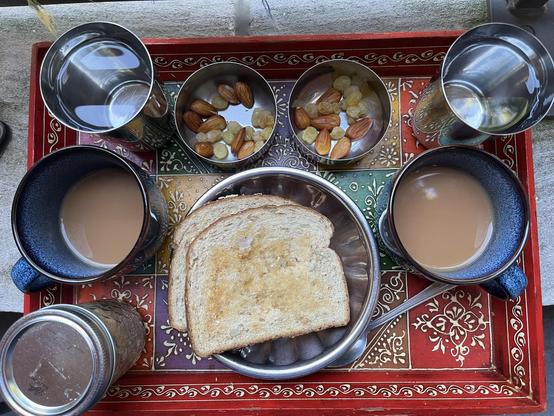A colorful tray with two stainless steel cups of water, two blue ceramic mugs of tea, two bowls of nuts and dried fruits, a few slices of toasted bread on a plate, and a jar.