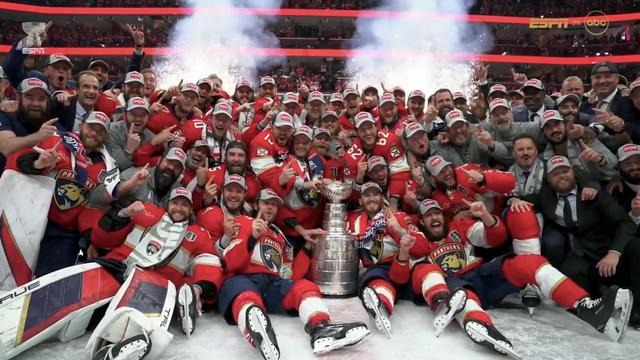 A hockey team celebrates on the ice, posing with the Stanley Cup. The players are wearing red jerseys and caps and are surrounded by fireworks.