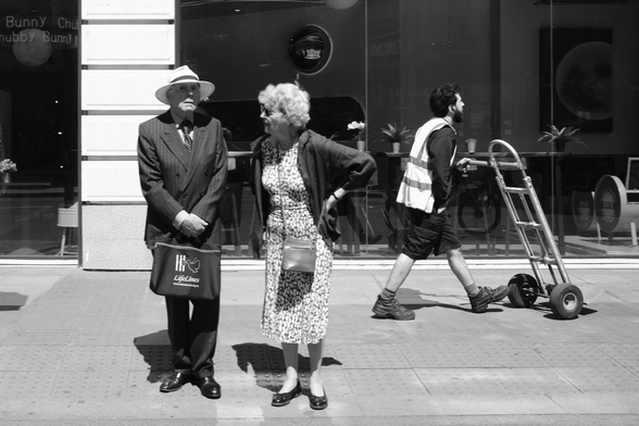 A couple stand on the street with a worker pushing a cart behind