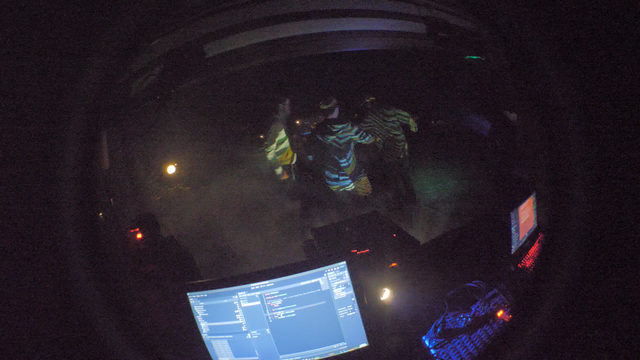 In the background a few people dancing  with colorful patterns being projected onto them from a short-throw projector.
In the foreground a control desk with a laptop running Godot editor, that is used for live modification of the projected visuals.