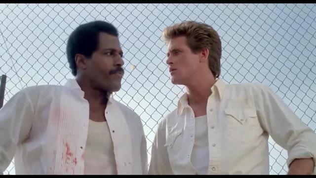 Two men stand in front of a chain-link fence, both wearing white shirts. One has blood stains on his shirt.