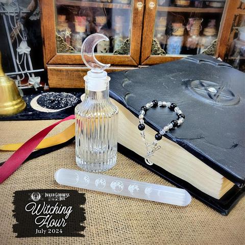 Three items are arrayed amongst props. The first is a bracelet made of round black, clear, and silver beads embossed with pentacles. Next, a clear fluted glass bottle has a silicone shank with a glass finial shaped like a crescent moon. Last, a cylindrical selenite wand has rounded ends and has seven moon phases etched along most of its length on one side. These are just three of the items in the exclusive July 2024 "Witching Hour" Inked Goddess Creations Box.