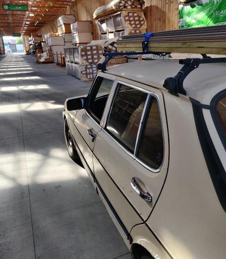 A beige, beater SAAB 900 with roof crossbars clamped to the gutter rails. The crossbars have large knobs on each end to screw tightly to the gutters. A small stack of lumber is strapped on top, and the car is parked in a large lumberyard shed and surrounded by large stacks of timber and plywood.
