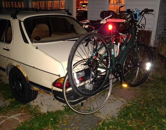 Same beige SAAB 900 at night, with two bikes strapped onto a flimsy-looking bike carrier on the trunk. The center of the carrier is raised so it does not contact the trailing edge of the trunk lid.