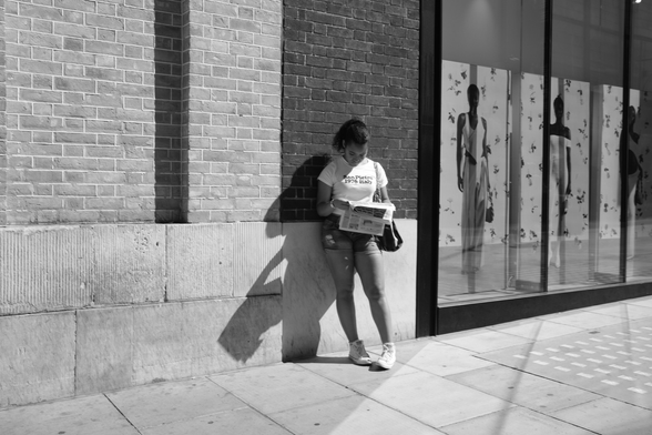 Woman reads newspaper against a wall.