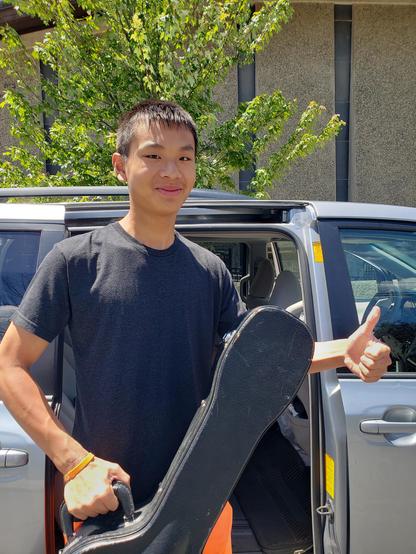 Student holding a guitar in a hard case. He is standing in front of a van, giving a thumbs up and smiling.