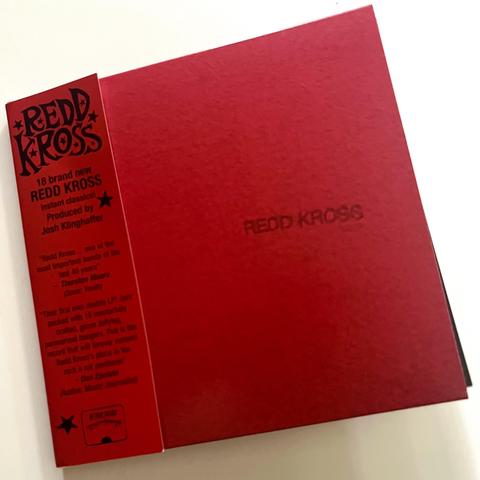 A red album cover with the text "REDD KROSS" in the center, accompanied by a vertical band with promotional text and quotes on the left side.