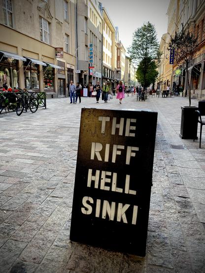 A street scene with shops, people walking, and a blackboard sign that reads "THE RIFF HELLSINKI" in the foreground.