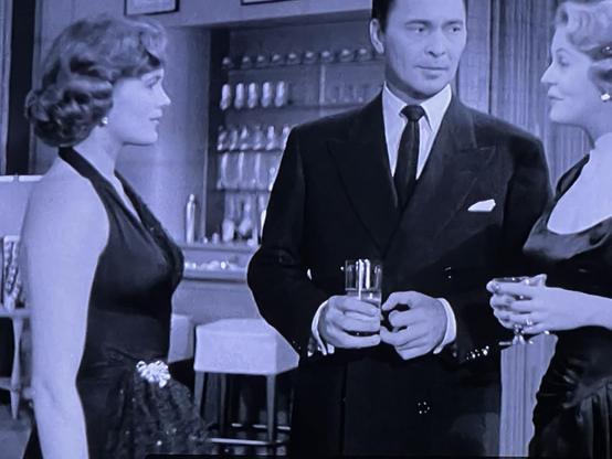 Three people in formal attire are conversing and holding drinks in a bar setting.