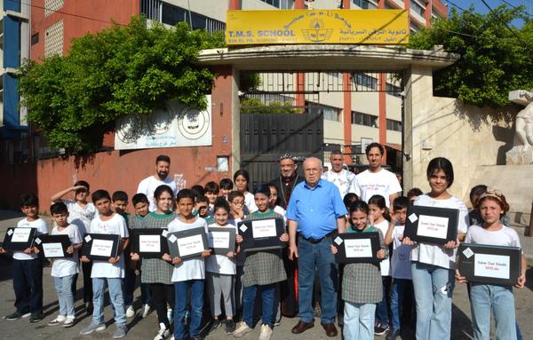 Vor einem Schulgebäude im Libanon steht eine Schülergruppe im Alter von ca. 8 bis 12 Jahren, mit Lehrern, Lehrerinnen und Flugpaten. Die Gruppe schaut lächelnd in die Kamera, einige Kinder halten Laptops in den Händen. Das Schulgebäude ist Ockerfarben gestrichen, einige grüne Büsche wachsen über eine Mauer.