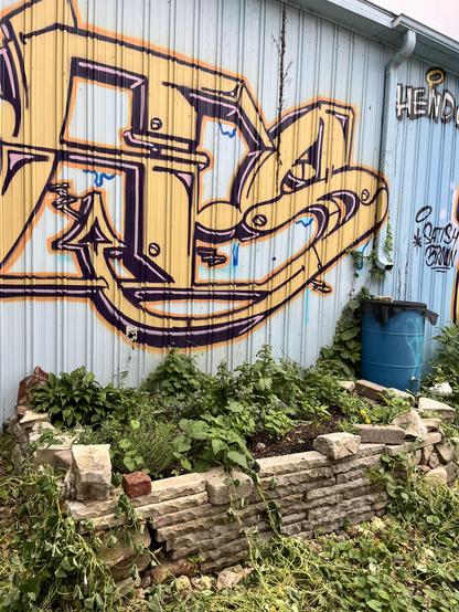 an aluminum siding shed with yellow graffiti on it; a raised stone wall possible garden is in front