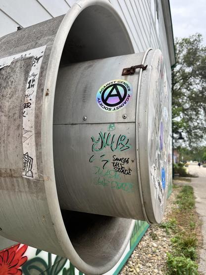 out of a wall sticker a metal tube with a second, closed, metal tube within it. There is some graffiti and an anarchy "A" sticker with the words "Against Gender; Against Society."