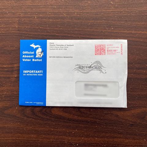 Photo of an "Official Absent Voter Ballot" on a table. Stylized icon of a map of Michigan in the top left, with a check mark superimposed. Return address is "Charter Township of Ypsilanti".