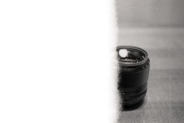 A scan of a 35mm black and white negative film with two-thirds of the photo completely overexposed to light (during the loading process).  The one-third properly exposed portion features just-over-half of a camera lens standing upright on a table, with a UV filter threaded onto it.  An overhead light is reflected in the filter's glass, causing an interesting glare of concentrated light.