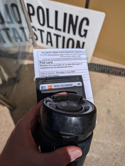Photo of a polling card held in front of a polling station sign. The card is partially covered by a wallet with a Spanish ID card on it and a reusable coffee cup.