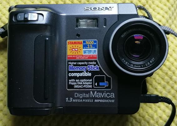 Vintage Sony floppy disk based digital camera. Square with two tone metallic grey. A large handle grip bulge is in the left and the lense in the right. The Sony brand in raised silver is above left if the lens, above the inset flash. In the middle of the flat fronted camera is a pristine metal stick in bright red, yellow and blue that details its technical features: stamina battery life, memory stick compatible, Digital Mavica, 1.3 megapixels, mpeg movie.