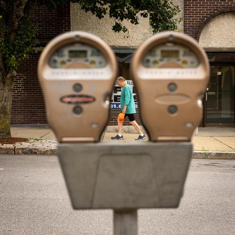 Color photo close up of a pair of parking meters which are out of focus. In the background, on the sidewalk across the street, a man walking is captured mid-stride right between them.