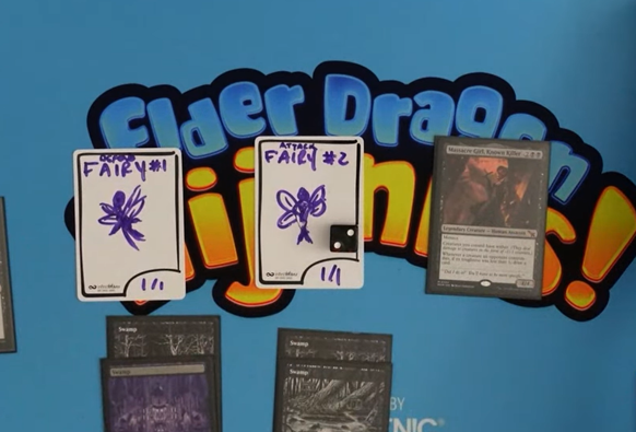 An Elder Dragon hijinks playmat with Massacre girl on the right and two InfiniToken dry erase tokens on the left. The dry erase tokens say "Attack Fairy" and "Defend Fairy" and have doodles of fairies.