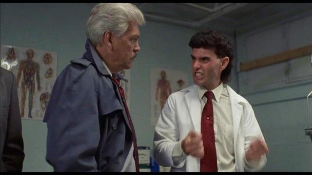 Elderly man in trench coat listens to middle-aged man in white lab coat and red tie who appears to be speaking passionately.