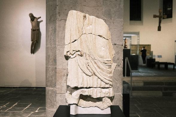 Fragment of a robed figure, late 10th cent. / 1st third 11th cent., shelly limestone, Cologne, St. Pantaleon. After the burial of Archbishop Bruno in the Church of St. Pantaleon in 965, the Benedictine abbey he had recently founded underwent extensive reconstruction, during which the remains of St. Maurinus were discovered. Some twenty years later, a gift from Empress Theophanu also brought the relics of St. Albinus to the Church of St. Pantaleon. The Albinus altar, near which the empress was buried in 991, occupied a central position in the western annex of the church. Perhaps as early as the end of the 10th century, but no later than the 1020s, the westwork was renovated. The facade of the build-ing was decorated with an elaborate, monumental sculptural programme, of which three special fragments are on display here. The figure of Christ occupied a central position in the three-storey structure. The larger-than-life head of his sculpture has survived. The fragment of a beardless head probably belongs to an angel. The fragment of the life-size robed figure of a male saint could have been one of the church’s patrons or even Albinus.