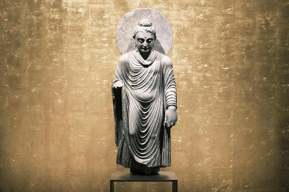 Standing Buddha, Pakistan, Gandhara, Takht-i-Bahi, 2nd -3rd c., schist, exhibited at the Humboldt Forum, Berlin. A deep sense of peace pervades the face of this standing Buddha. His eyes are lowered, and the facial features are completely smooth, free from any passion. The left hand hangs down, clasping a small loop of his robe, and the right arm is raised. The missing hand was probably raised in the gesture of protection (abhaya mudra).