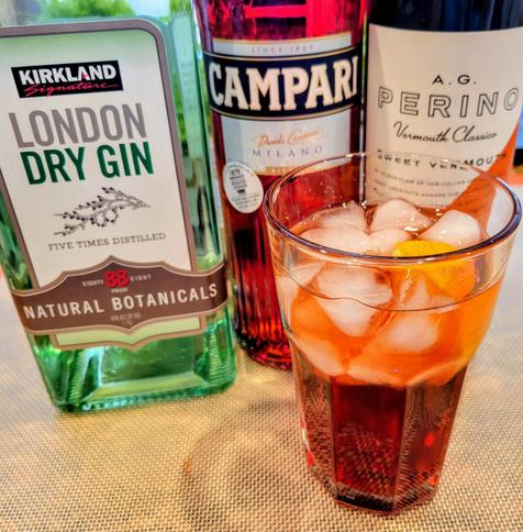 Photo of a Gibraltar glass with Negroni cocktail, in front of three bottles: 1) Gin (Kirkland Signature London Dry Gin); 2) Campari; and 3) Sweet Vermouth (A.G. Perino Vermouth Classico). The glass has ice & an orange slice. They all rest on a silicone baking sheet.