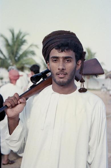 A young man wearing a white dishdasha and turban and carrying a rifle.