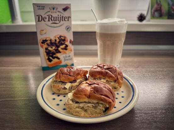 A plate with three Dutch raisin breadrolls, behind the plate a box of Dutch chocolate crumble „De Ruijter vlokfeest“ and a latte macchiato. 