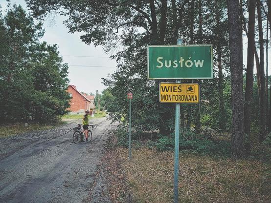Leśna droga prowadząca do zabudowań. Przy drodze tablice informacyjne. Pierwsza z napisem Susłów. Druga, poniżej pierwszej, z napisem: wieś monitorowana.