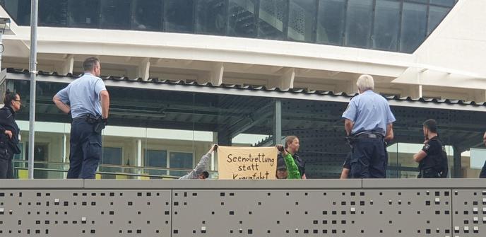 Aktivisti halten ein Banner mit der Aufschrift "Seenotrettung statt Kreuzfahrt", um sie herum stehen Polizist*innen.