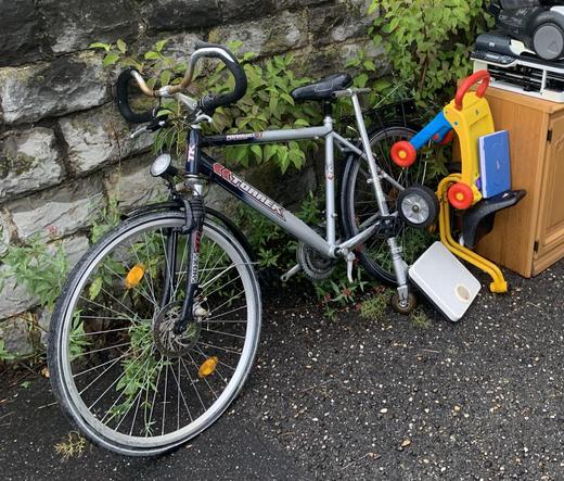 A silver bicycle leaning against a stone wall, surrounded by miscellaneous items including a toy bike with training wheels, a small blue book, and a wooden cabinet.