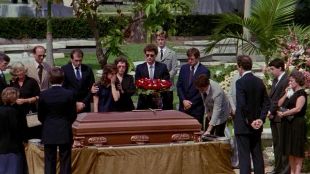 A group of people gathered around a coffin at a funeral in an outdoor setting. Some are visibly mourning while others stand solemnly.