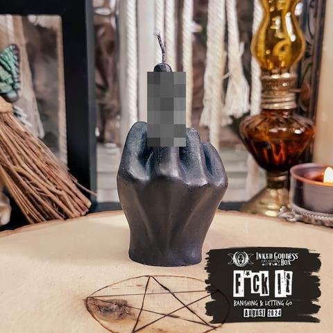 A black candle sits on a raw wood slice. The candle is shaped like a human hand from the wrist, held in a fist. Its middle finger is extended, but we have to pretend we don't know what it is because it has a censored pixel effect on the finger. This is one of the items in the exclusive August 2024 "F*ck It: Banishing & Letting Go" Inked Goddess Creations Box.