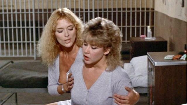 Two women in a room with a bed, one sitting and the other standing behind her, holding her shoulders. The background shows a barred window or door, suggesting a prison or similar facility.