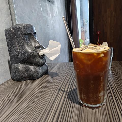 The same iced coffee, but there is a tissue dispenser in the background. The tissue dispenser is in the shape of a statue's head, very similar to the ones on Easter Island. The tissues get pulled from its round mouth, which gives the effect of the statue blowing them out.