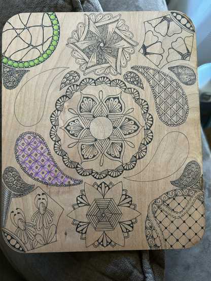 Wooden board with the start of a design on, mostly still black but some green coloured balls and some purple grid work 