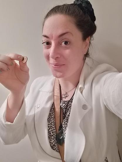 Colour; portrait orientation: Selfie of a Caucasian woman (me) smiling, with chipped metallic nail polish, dark hair tied back and embellished with a black faux flower, a tan bag strap slung over her shoulder, and wearing a white jacket, silver Eye Of Ra pendant, brown boulder opal earrings, a Maori pounamu pikorua pendant, and a leopard print T-shirt, holding a copper, 1989 dated Australian one cent coin.