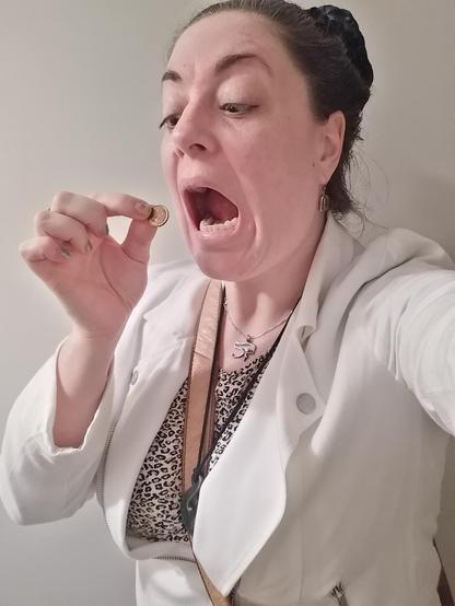 Colour; portrait orientation: Selfie of a Caucasian woman (me) being a goofball, with chipped metallic nail polish, dark hair tied back and embellished with a black faux flower, a tan bag strap slung over her shoulder, and wearing a white jacket, silver Eye Of Ra pendant, brown boulder opal earrings, a Maori pounamu pikorua pendant, and a leopard print T-shirt, pretending to be about to eat a copper, 1989 dated Australian one cent coin.